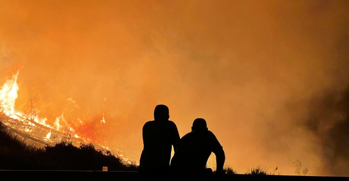 Silhouetted figures observe a large wildfire engulfing the landscape filling the sky with orange flames and smoke